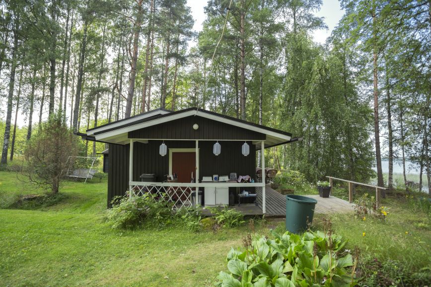 En liten mörkbrun stuga vid stranden av en sjö i ett frodigt landskap.